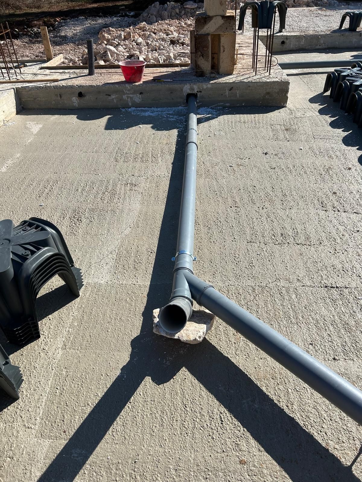 Tubo grigio rotto su un terreno di cemento accanto a una carriola e attrezzature nere all'aperto.