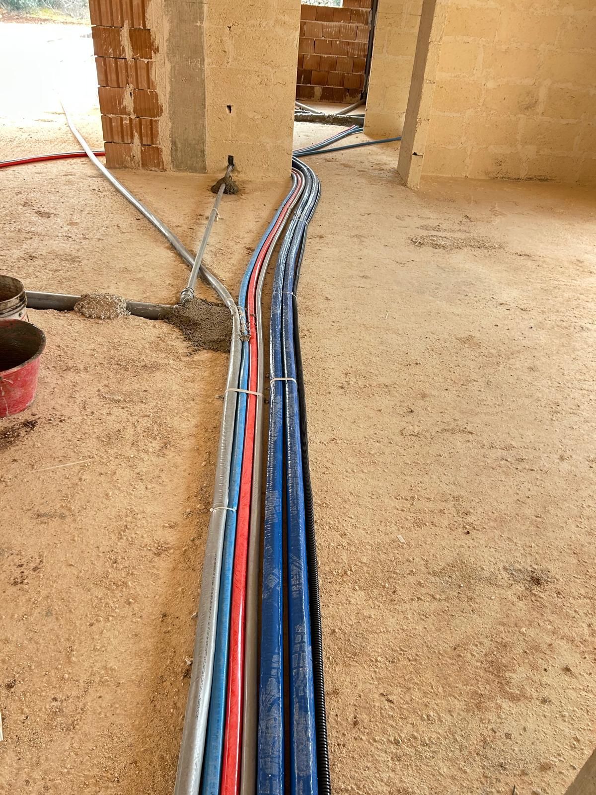 In un cantiere, dei cavi rossi, blu e neri attraversano un pavimento di terra battuta e passano attraverso una porta.