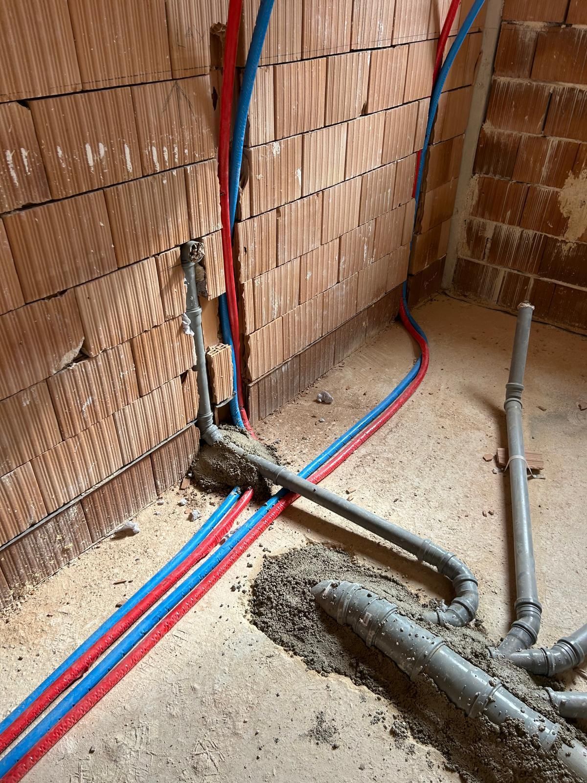 Muro di cantiere con tubature rosse e blu a vista in una trincea rivestita di mattoni, circondato da polvere e detriti.