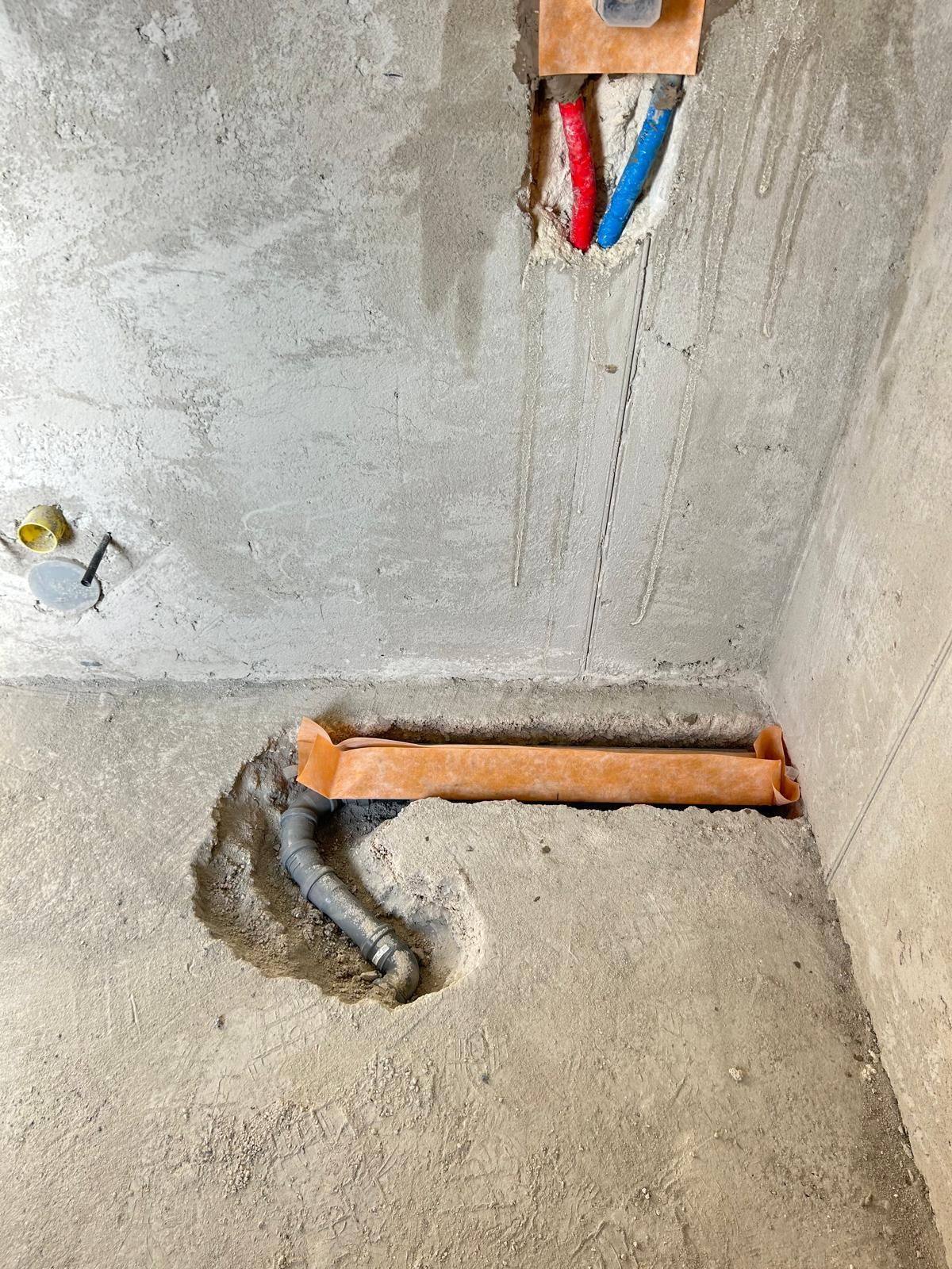 Fili elettrici scoperti e un moncone di tubo di rame che spuntano da un angolo grezzo tra muro e pavimento in cemento.