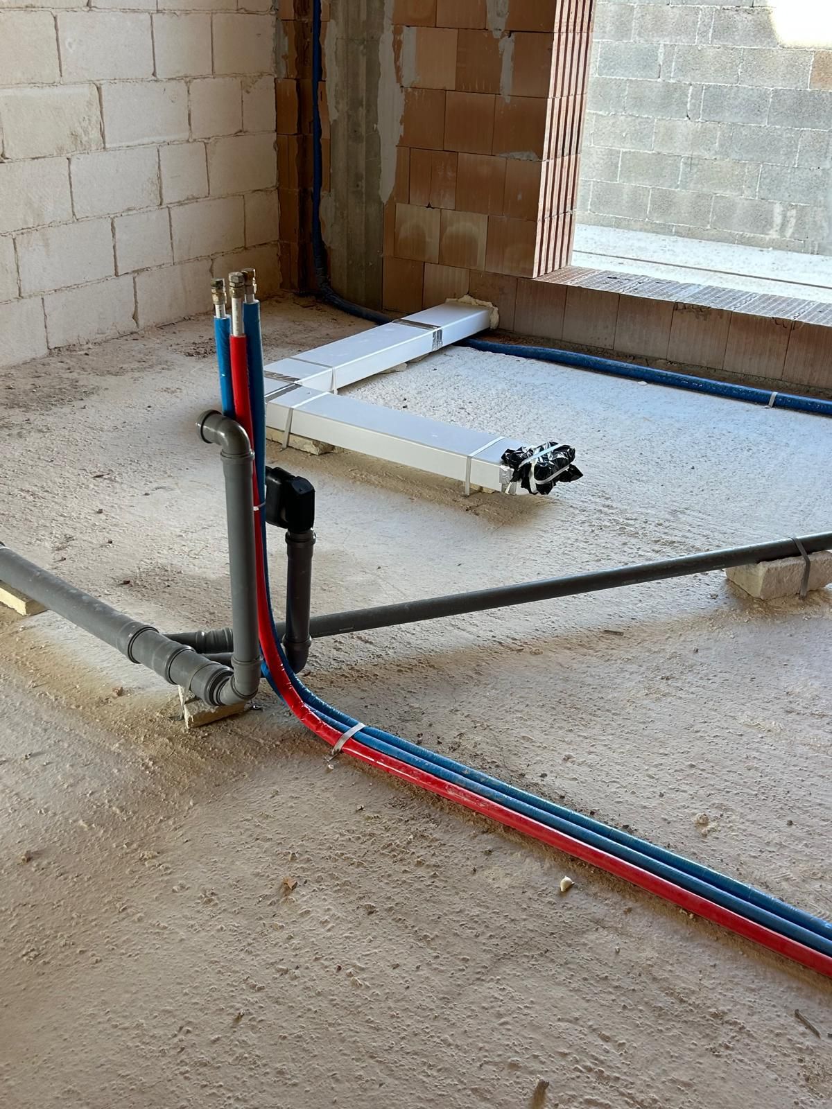 Pavimento di cantiere con tubature rosse e blu a vista accanto a una parete e a un'apertura per una finestra.