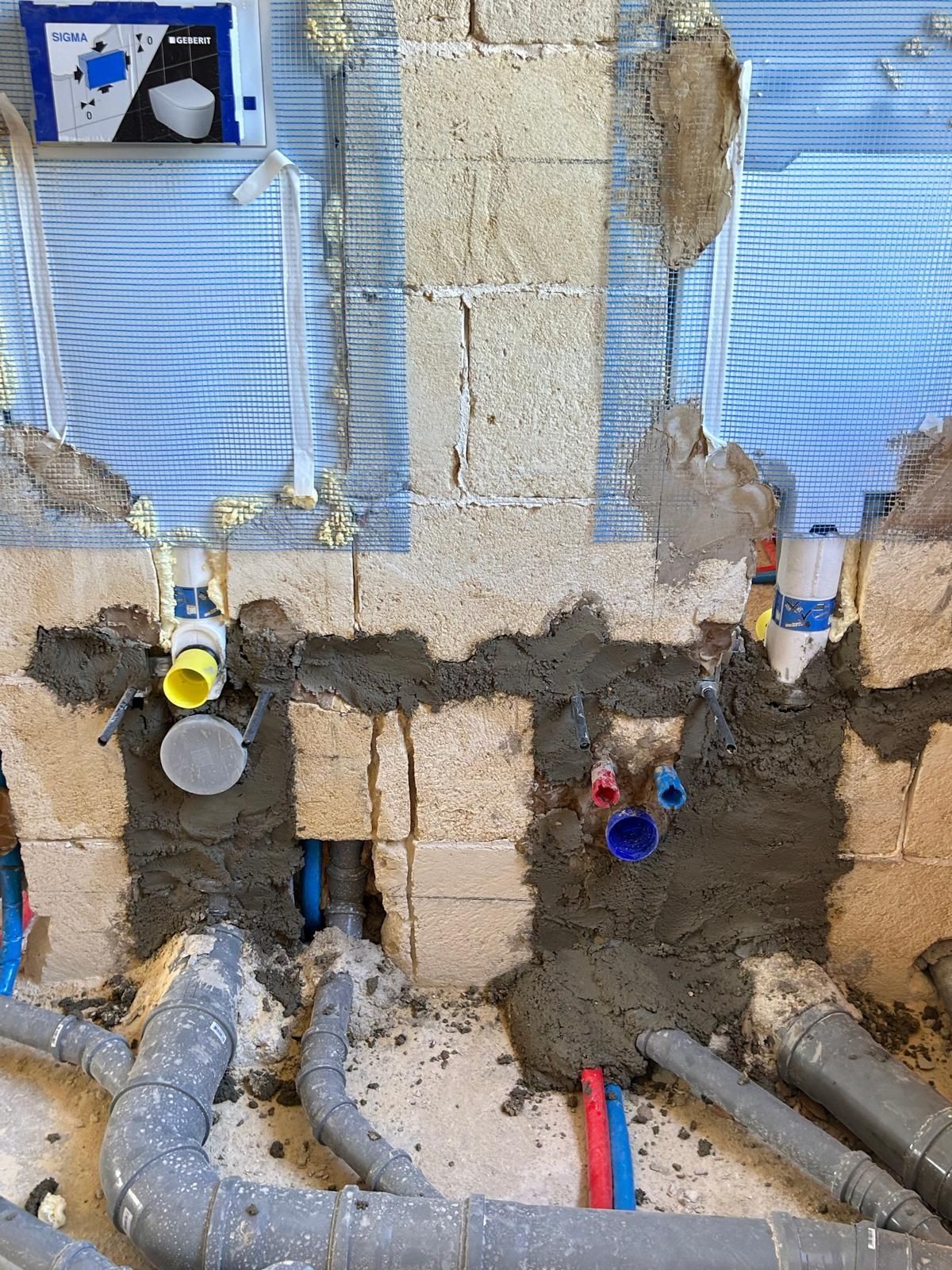 Tubature e condotte a vista in un muro parzialmente demolito, con isolamento blu e riparazioni in cemento grezzo.