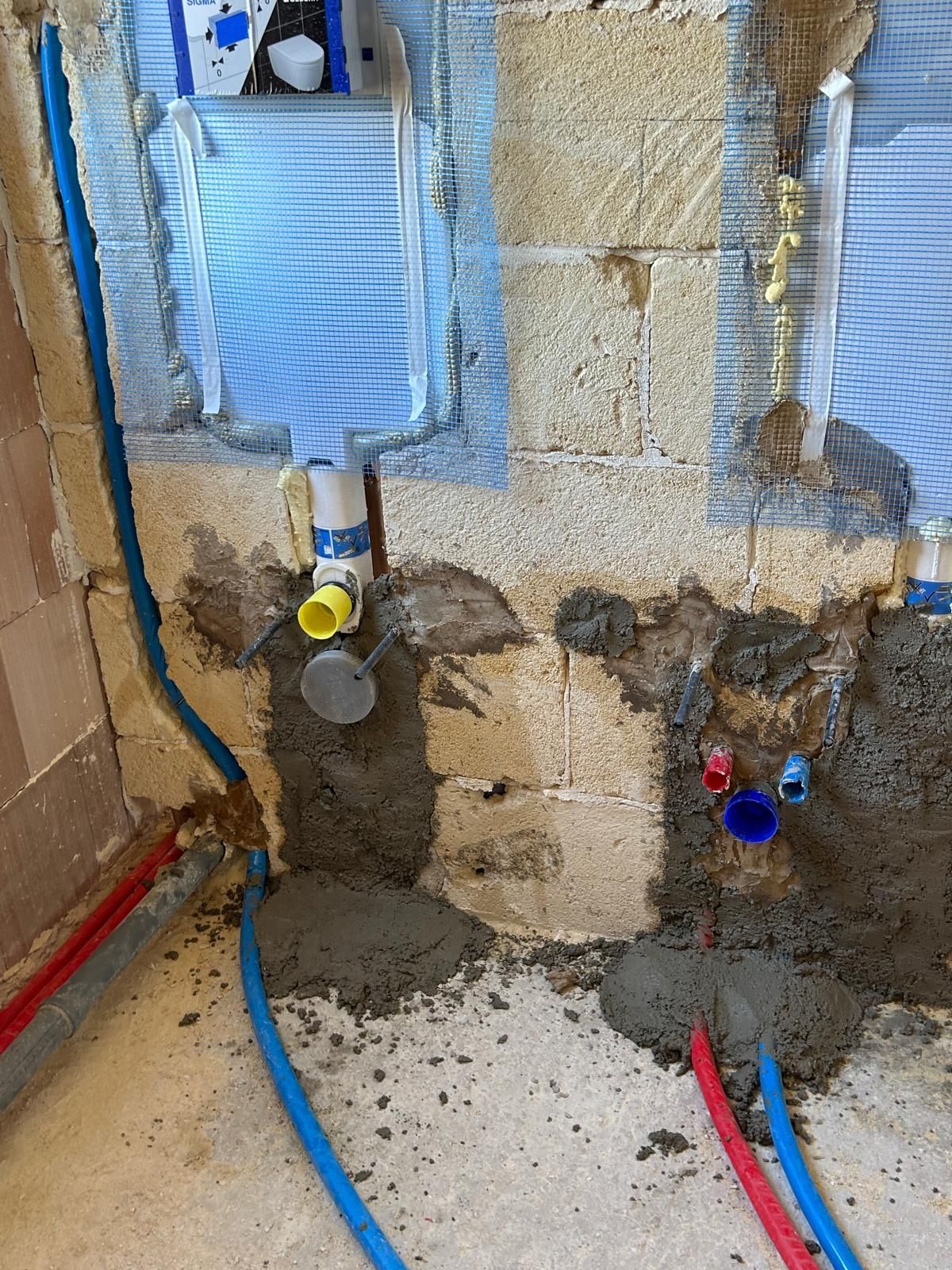 Predisposizione idraulica con parete a vista, tubi blu e rossi e scarichi a pavimento in un cantiere.