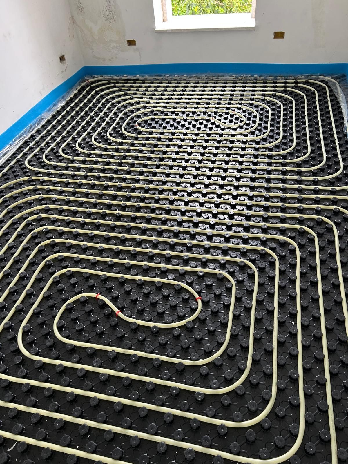 Tubi neri a spirale per il riscaldamento a pavimento, posati sopra l'isolamento in una stanza non rifinita con pareti bianche e una finestra.