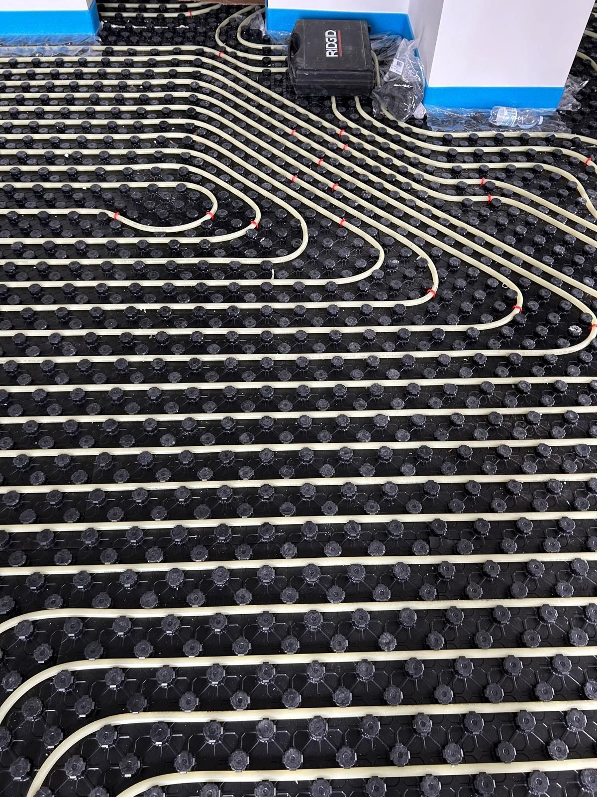 Sottotappeto nero con tubi di riscaldamento bianchi e una piccola scatola elettrica vicino a una parete