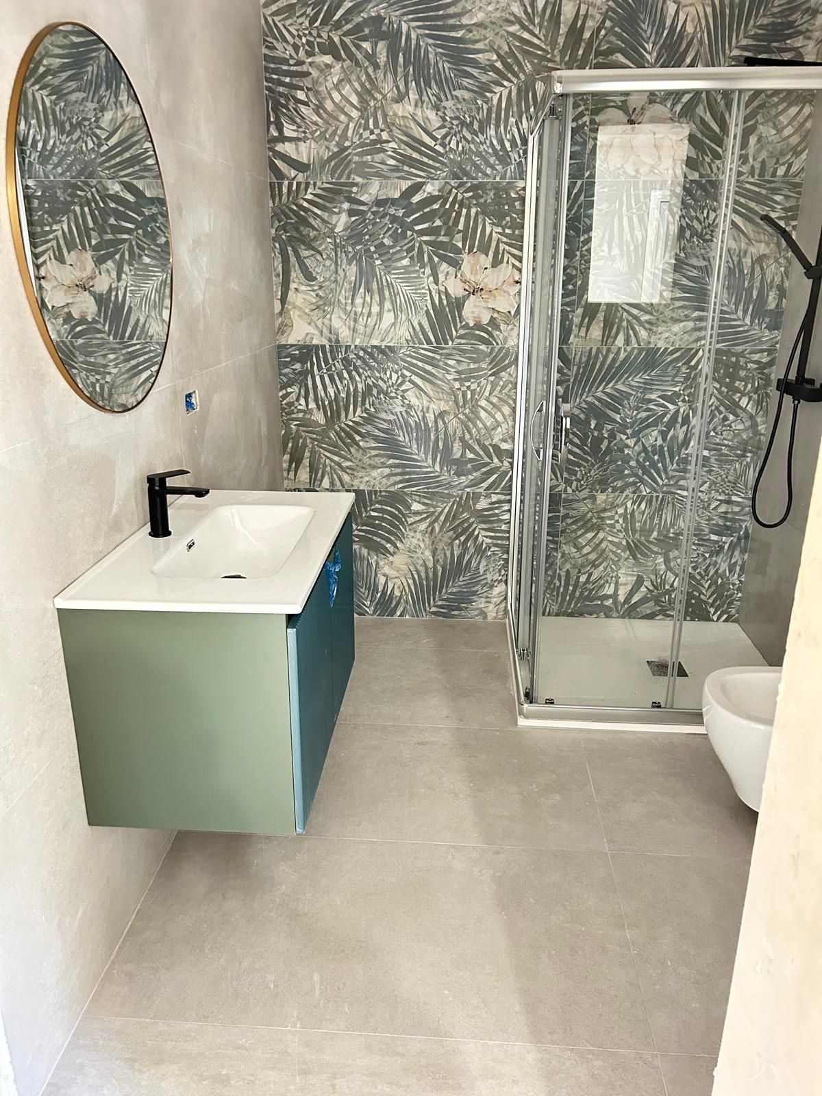 Bagno moderno con carta da parati tropicale, specchio rotondo, mobile lavabo sospeso, doccia in vetro e WC.