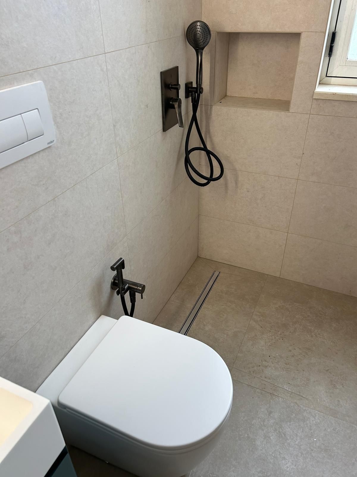 Piccolo bagno beige con WC, bidet portatile, doccetta e pavimento piastrellato.