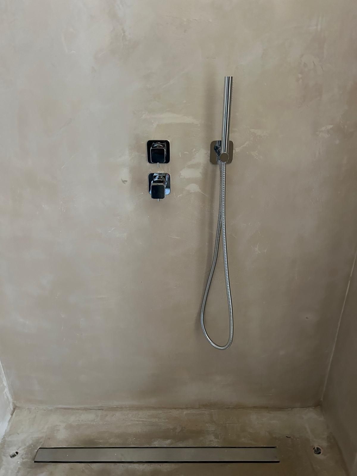 Box doccia minimalista con doccetta a mano a parete, comandi e scarico a pavimento.