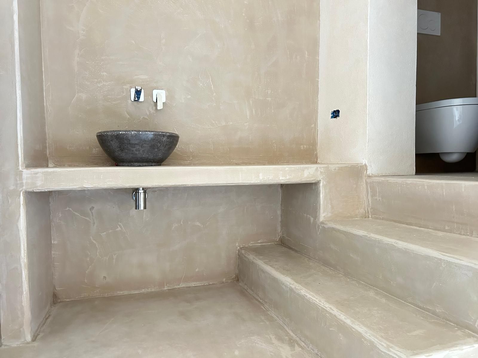 Bagno minimalista sui toni del beige con lavabo in pietra e scalini integrati che conducono alla zona vasca.