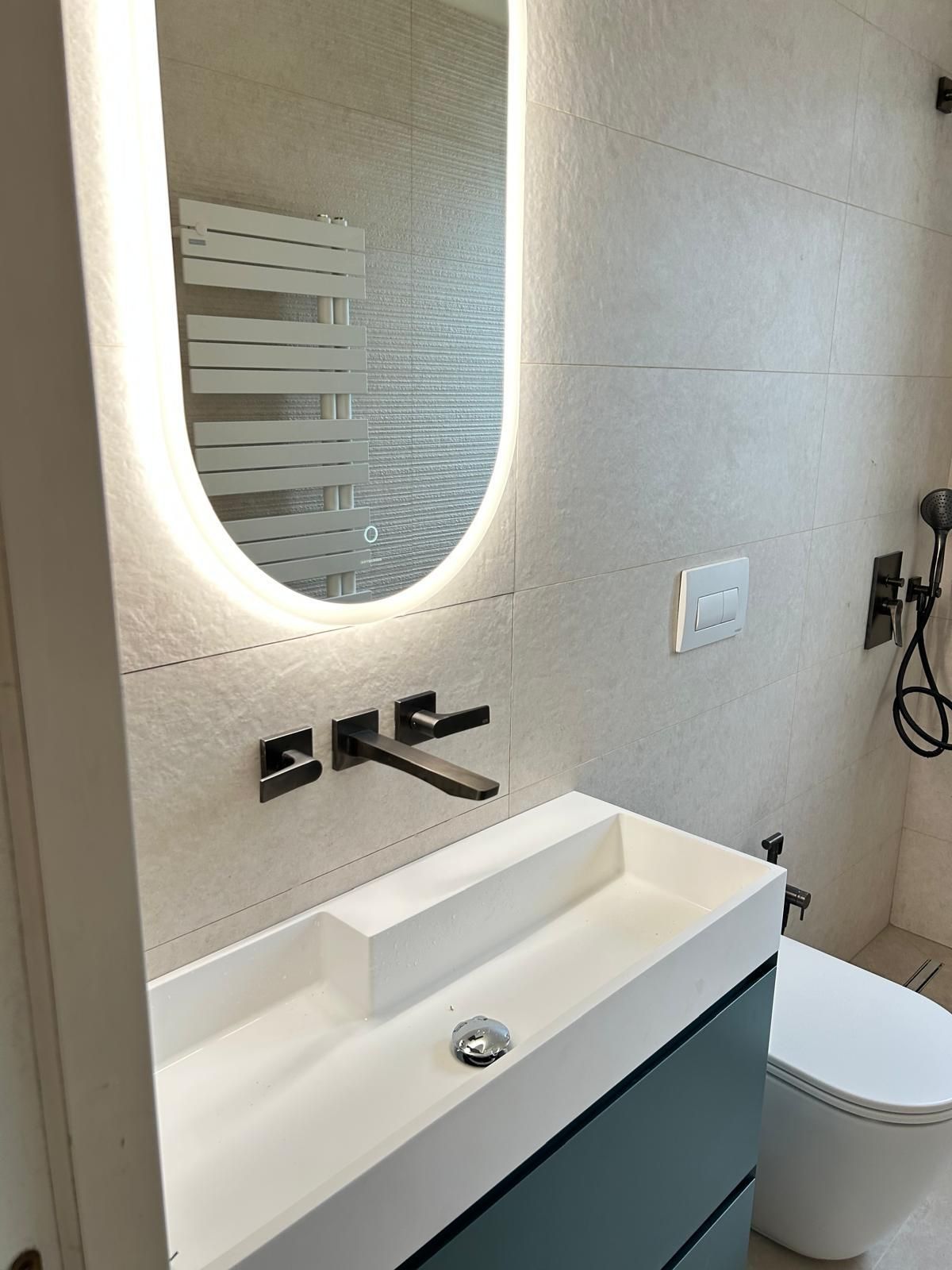 Mobile bagno moderno con specchio ovale illuminato, rubinetto nero e WC accanto a una cabina doccia.