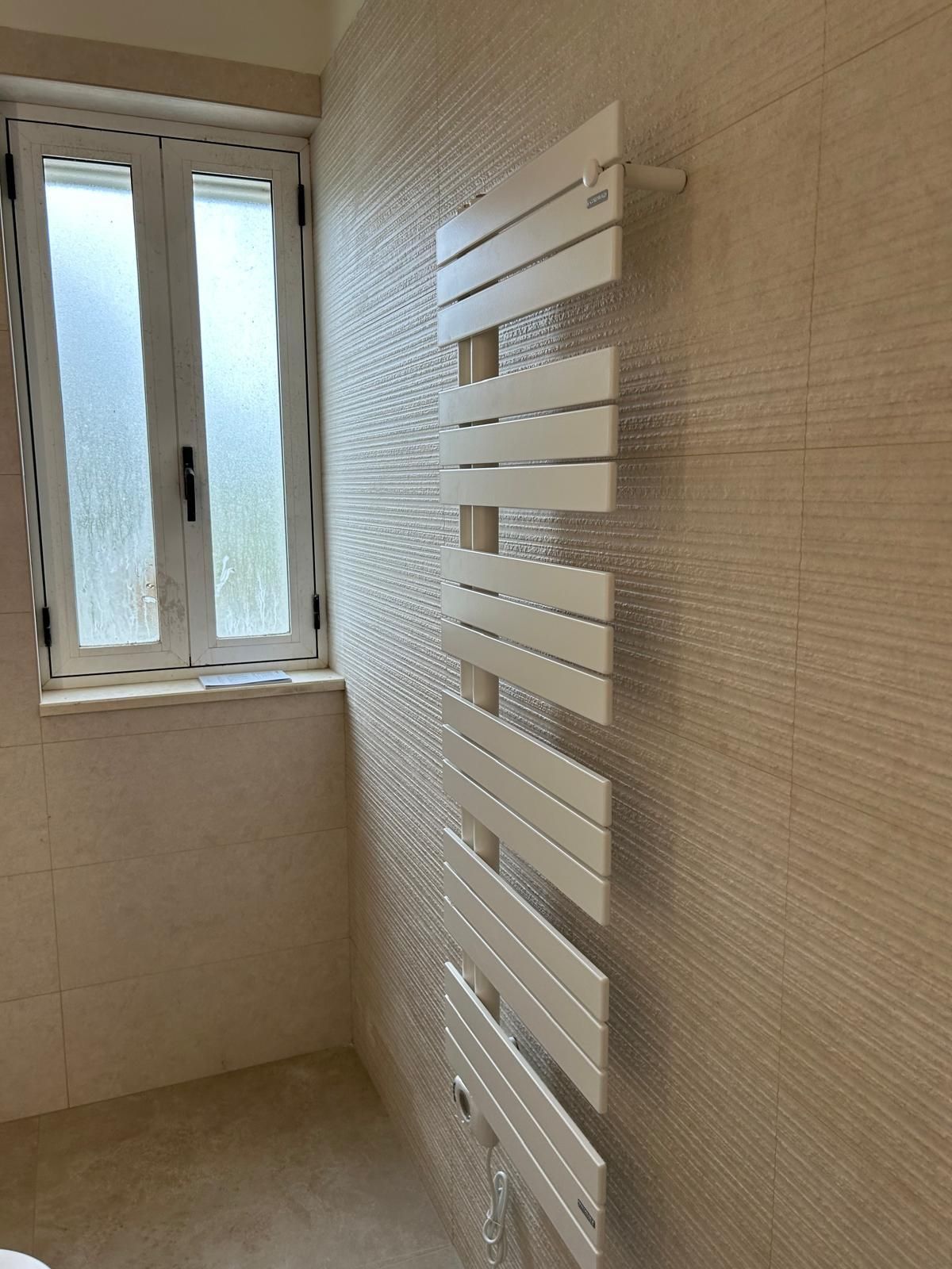 Piccolo bagno con termoarredo bianco accanto a una finestra e pareti piastrellate.