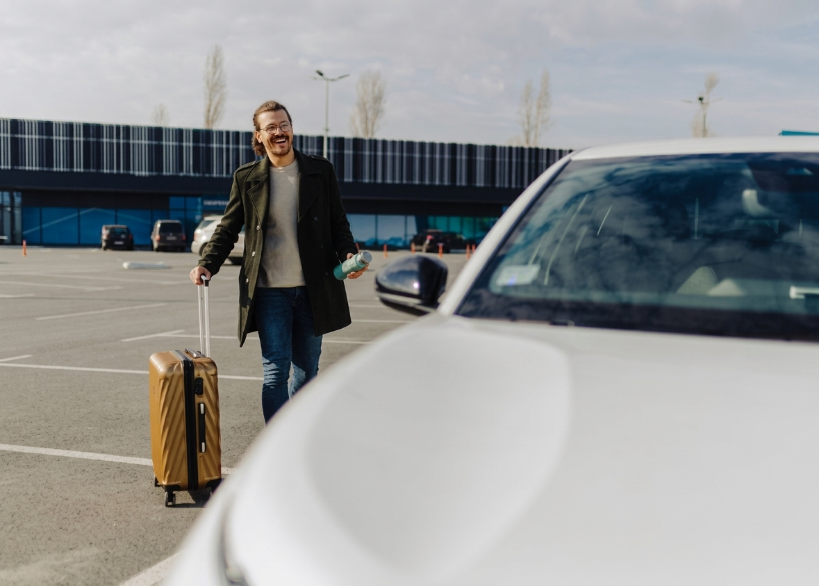 Un viaggiatore cammina accanto a un'auto bianca in un parcheggio dell'aeroporto con una valigia gialla.
