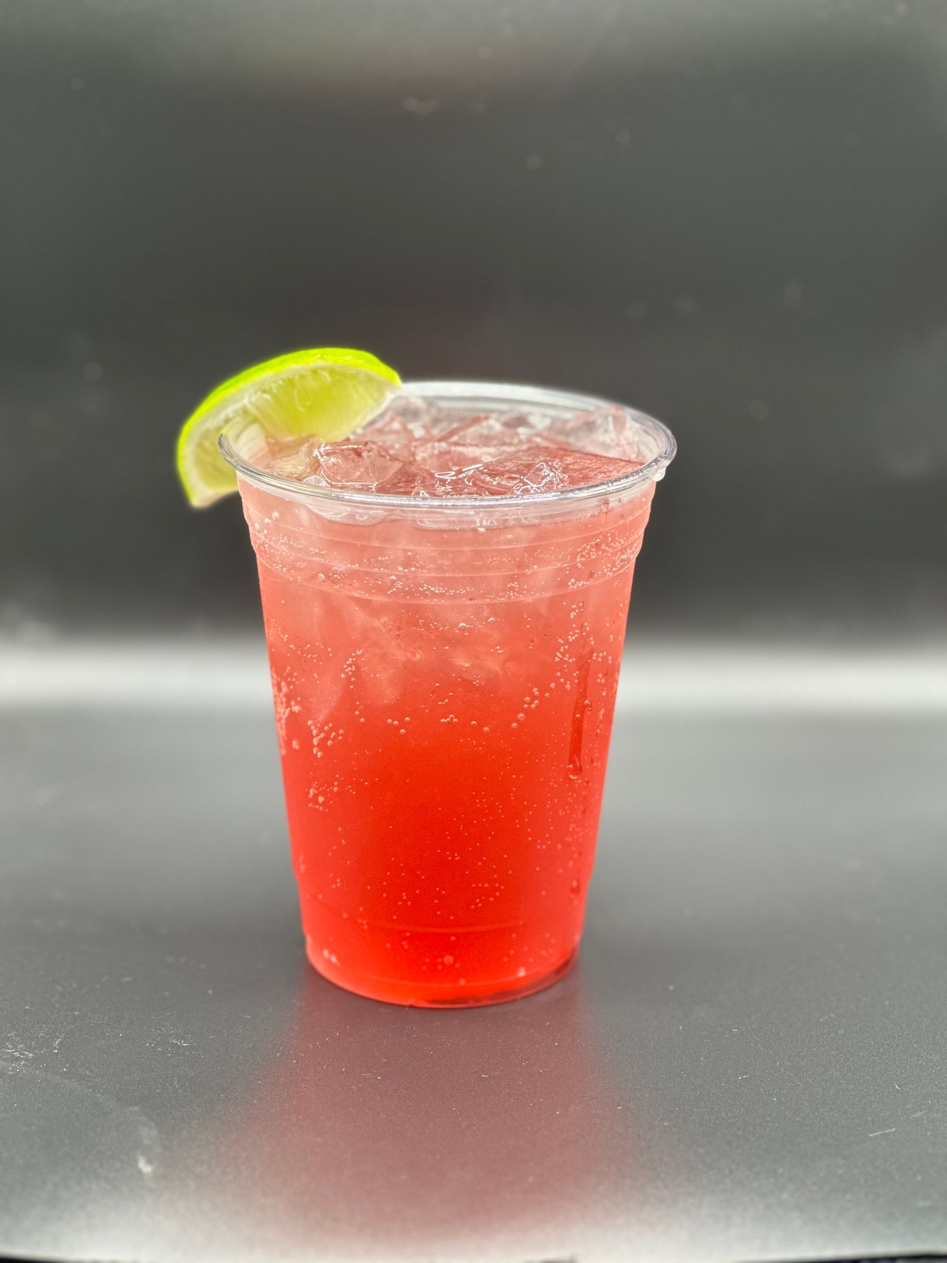 A red drink in a clear plastic cup, garnished with a lime wedge on a gray surface.