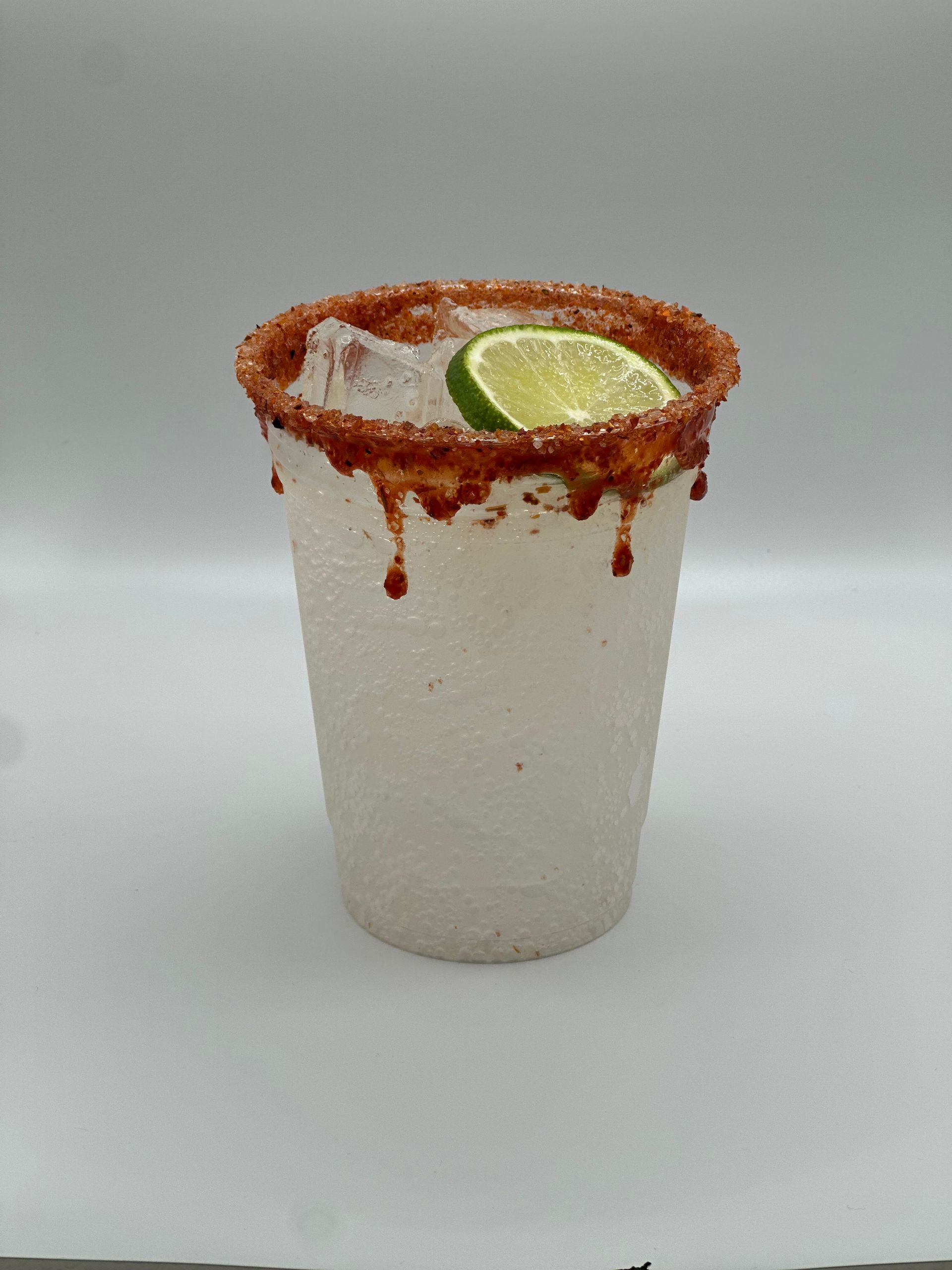 A clear plastic cup with a brown chili-salt rim, ice, and a lime slice, against a white background.