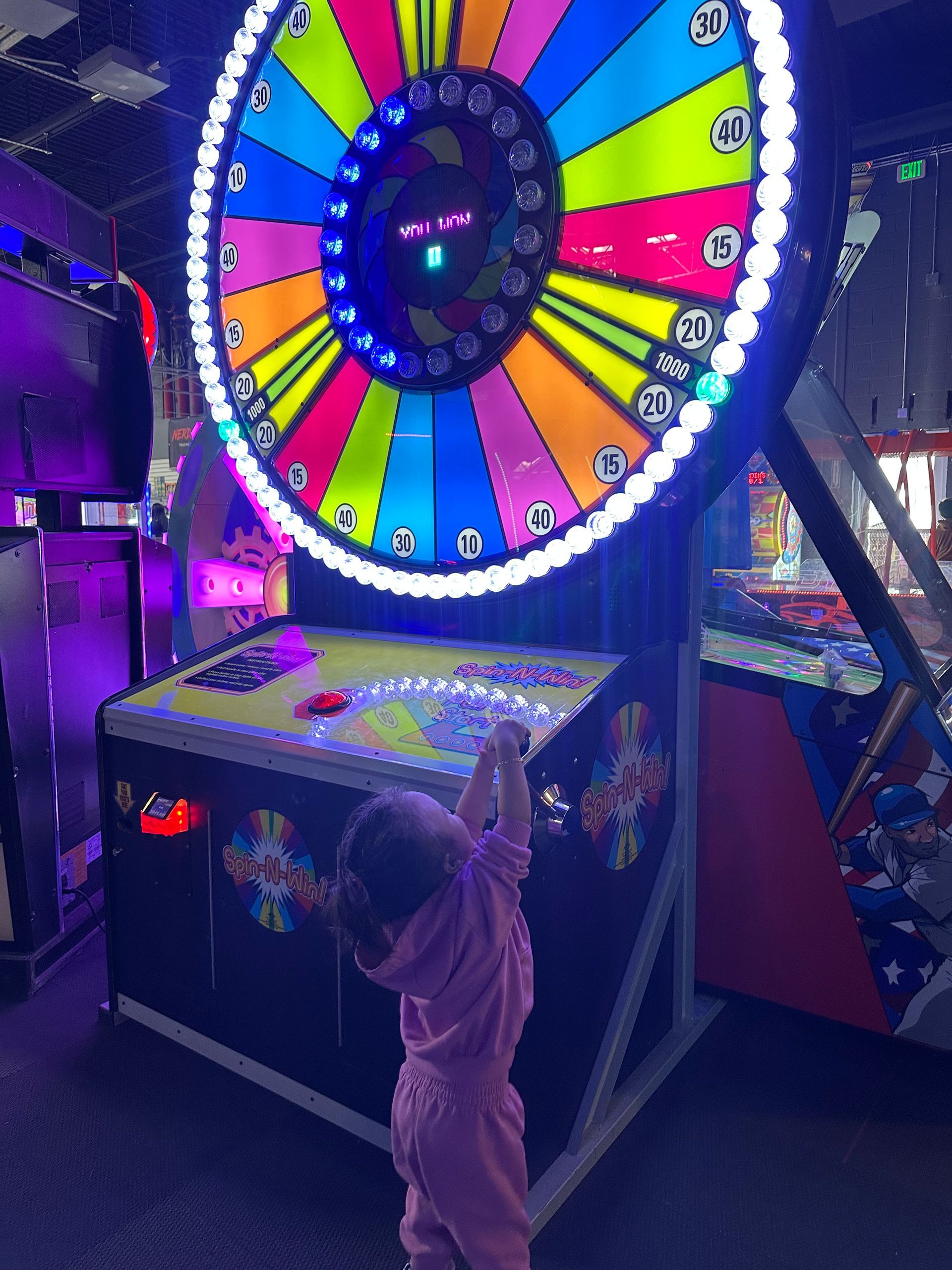 A young child in pink reaches up toward a brightly lit prize wheel game in an arcade.