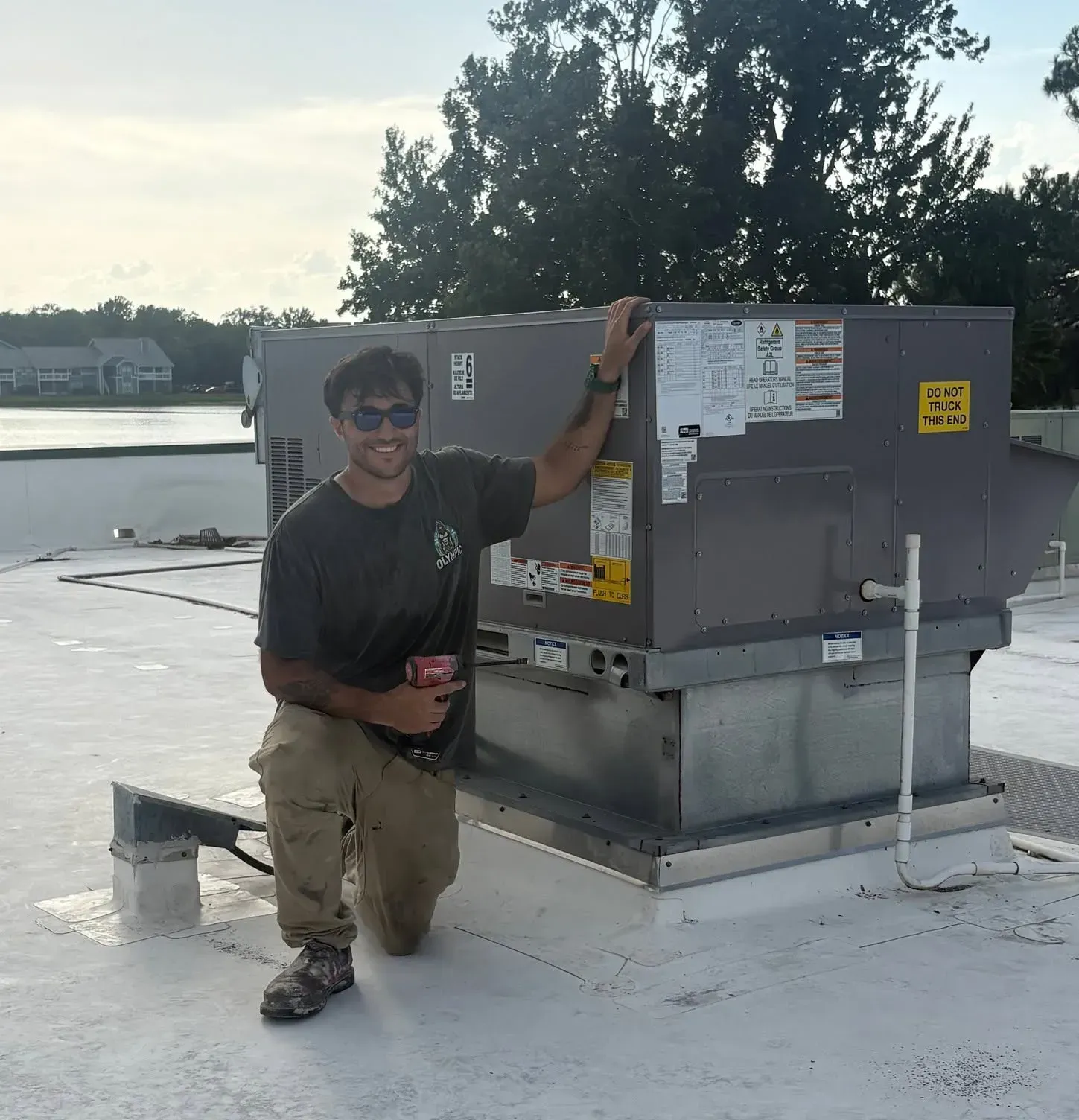 Person kneeling beside a gray rooftop HVAC unit by the water, wearing sunglasses and a dark shirt.