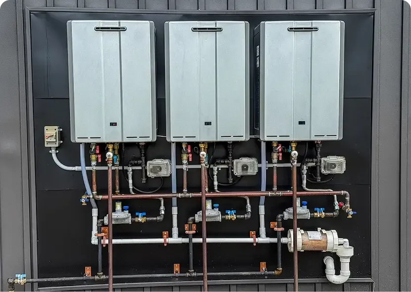 Three gray wall-mounted boilers with exposed copper piping and valves on a utility wall