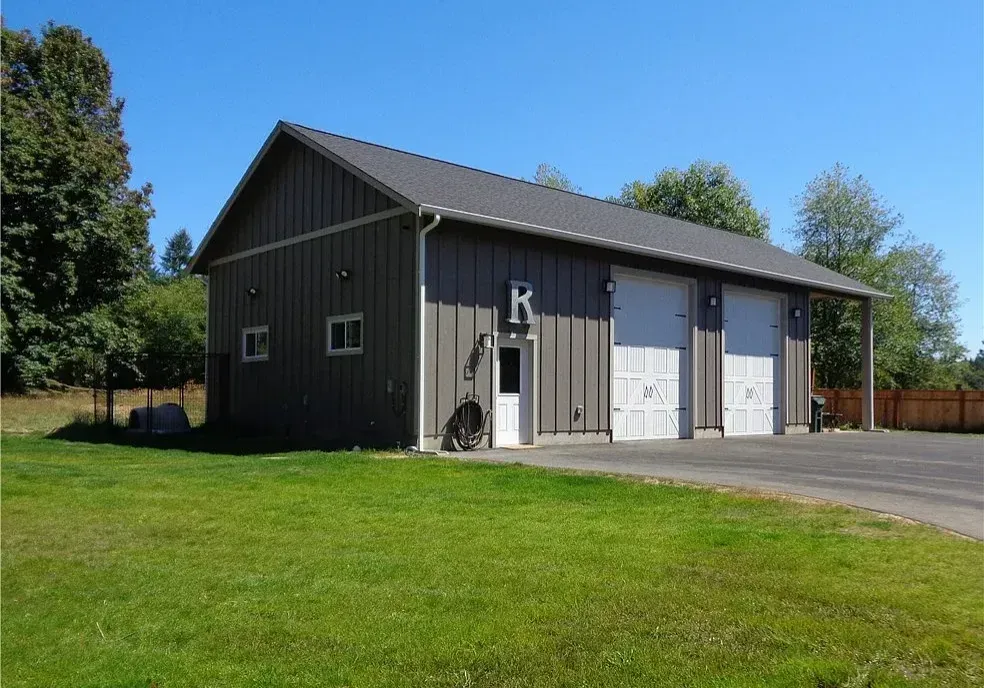 Gray metal garage building with three white doors beside a grassy lawn under a clear blue sky