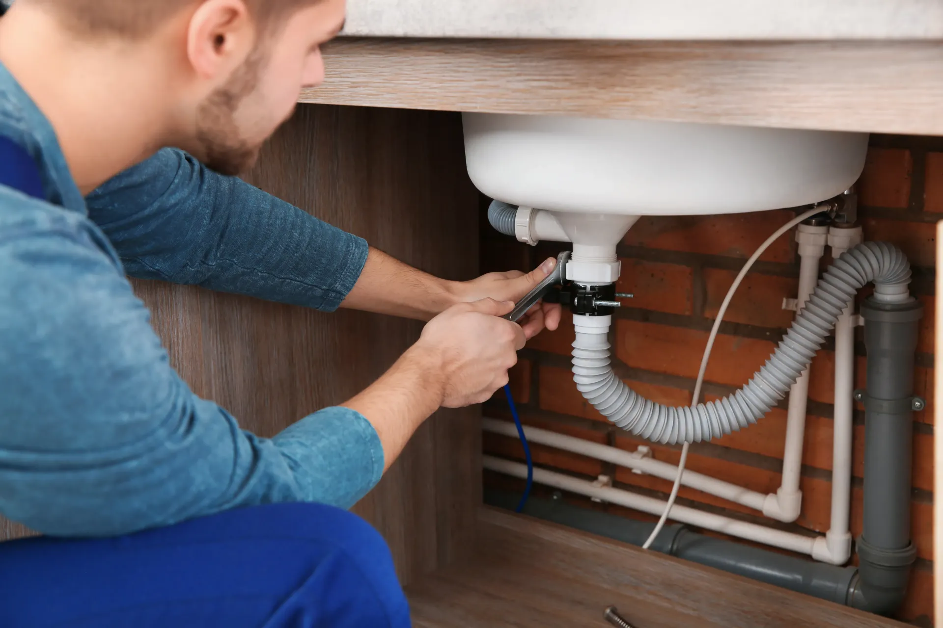 Person tightening pipes under a sink with a wrench in a wooden cabinet.