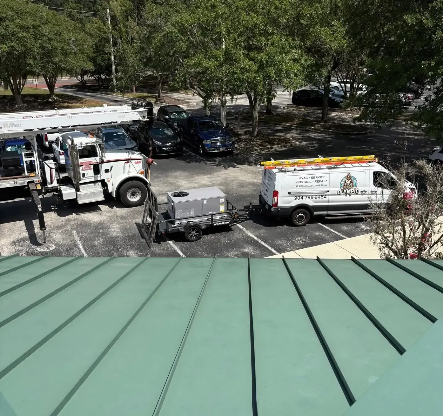 Tow truck and utility van parked by a green roof, with trees and parked cars in the background