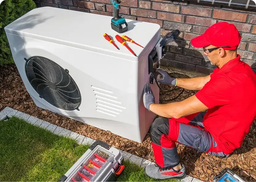 Technician in red uniform servicing an outdoor HVAC unit with tools beside a brick wall.