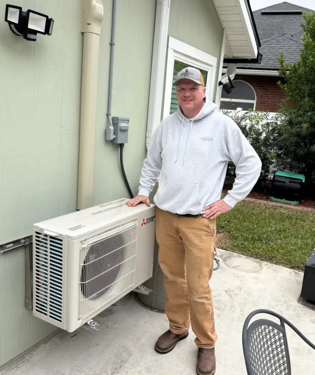 Man standing beside a wall-mounted outdoor AC unit next to a house exterior.