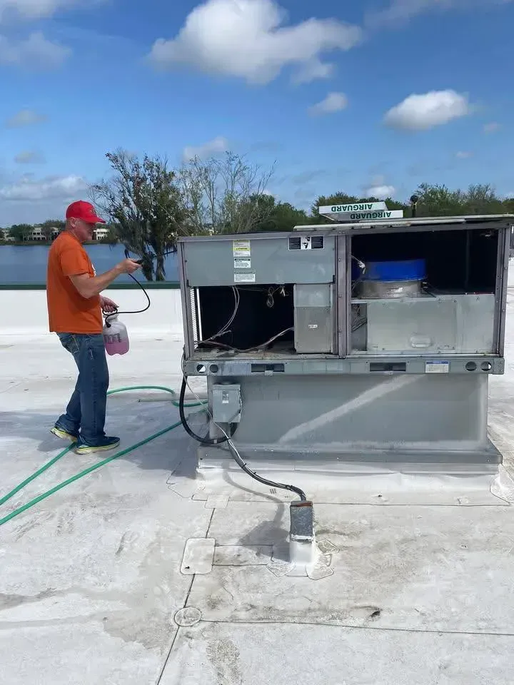 Worker in orange shirt and red hard hat inspects rooftop HVAC equipment under a clear sky.