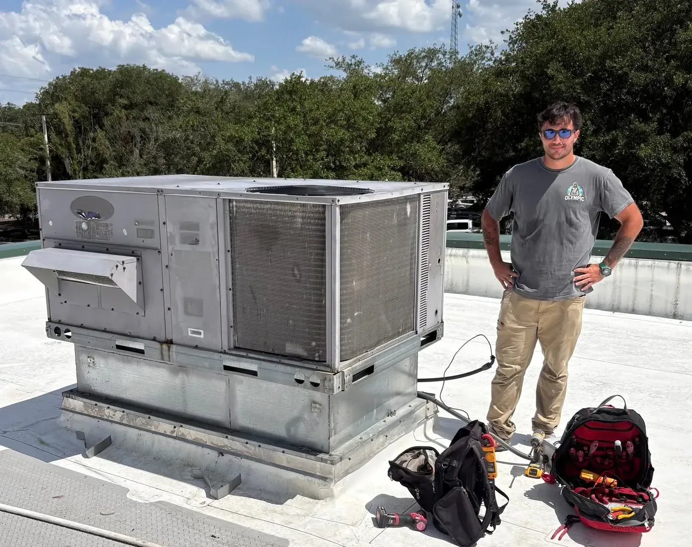 Man standing beside rooftop HVAC unit and tools on a flat roof under a partly cloudy sky