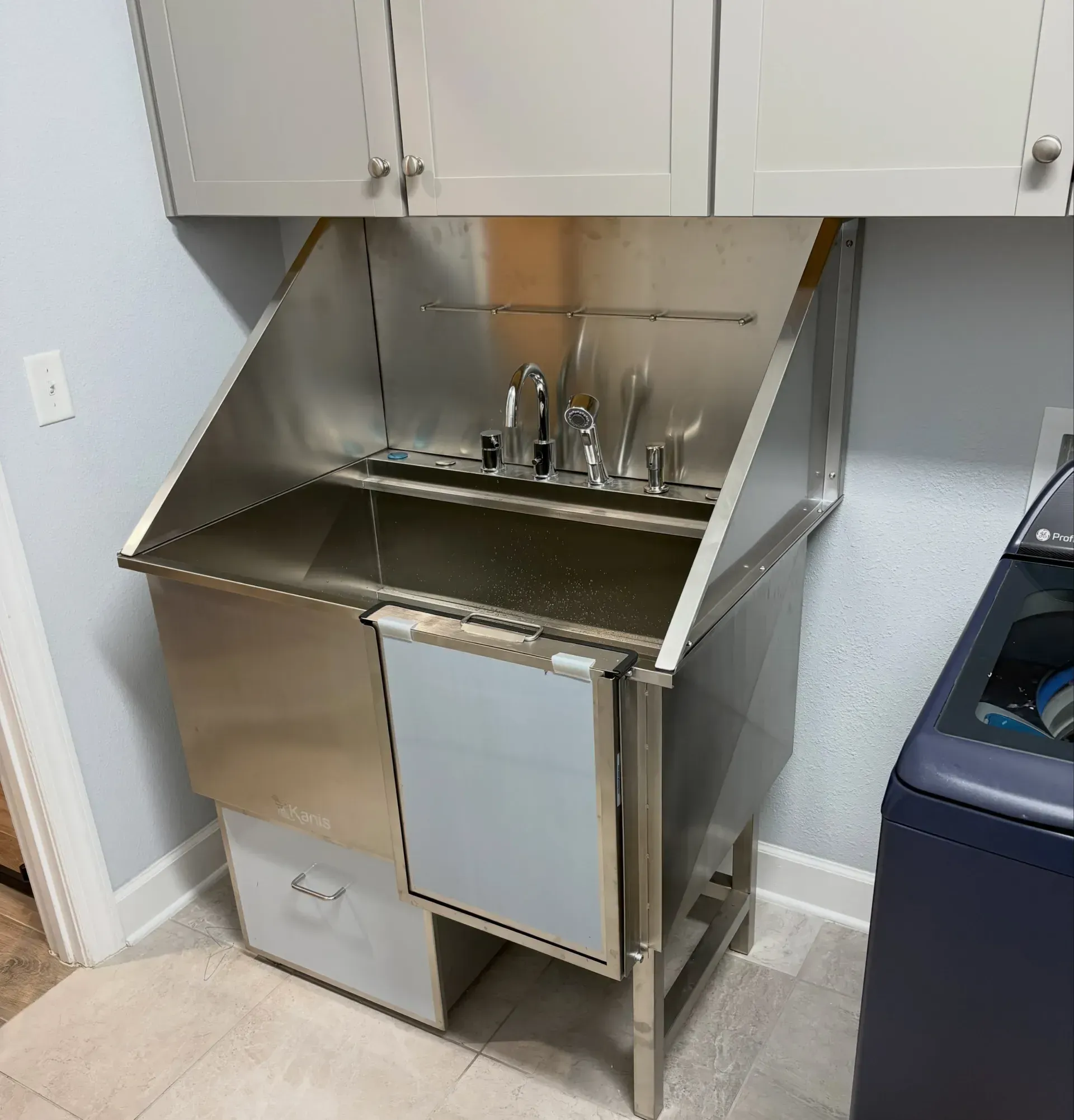 Stainless-steel utility sink with cabinets above, a faucet, and a white panel in a small laundry or utility room.