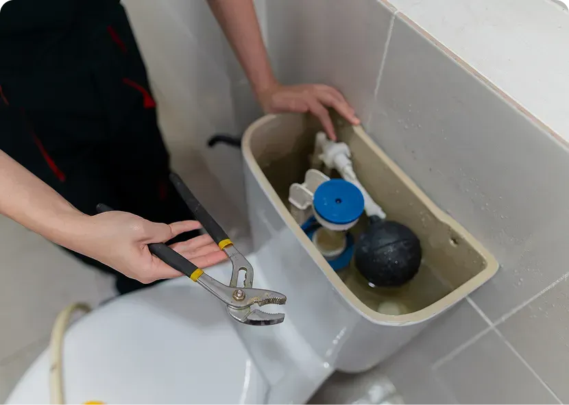 Person using pliers to adjust toilet tank parts with water inside, next to a bathroom sink.