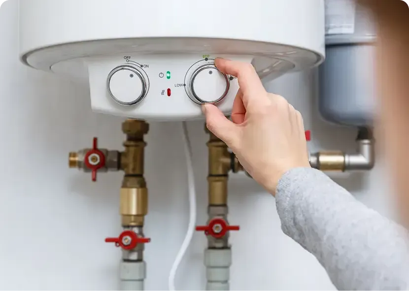 Hand adjusting a white water heater’s temperature dial above exposed pipes and red valves