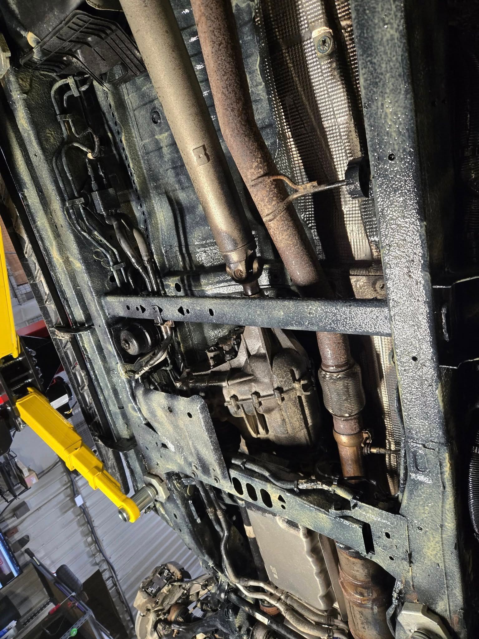 Underside of a vehicle with rusted exhaust pipes and a yellow lift arm in a repair garage