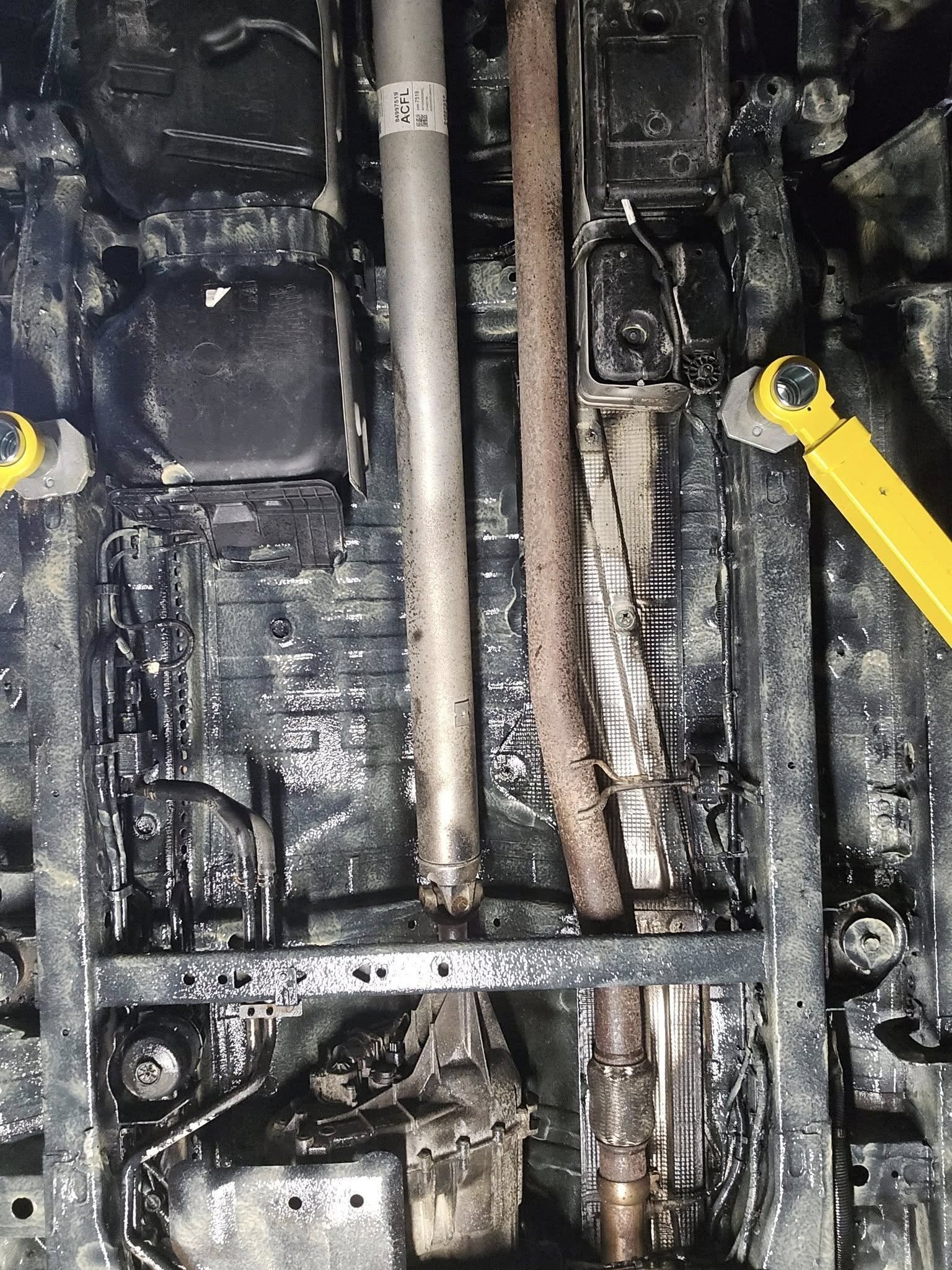 Dirty mechanical compartment with metal pipes, chains, and yellow hoses inside a dark engine bay.