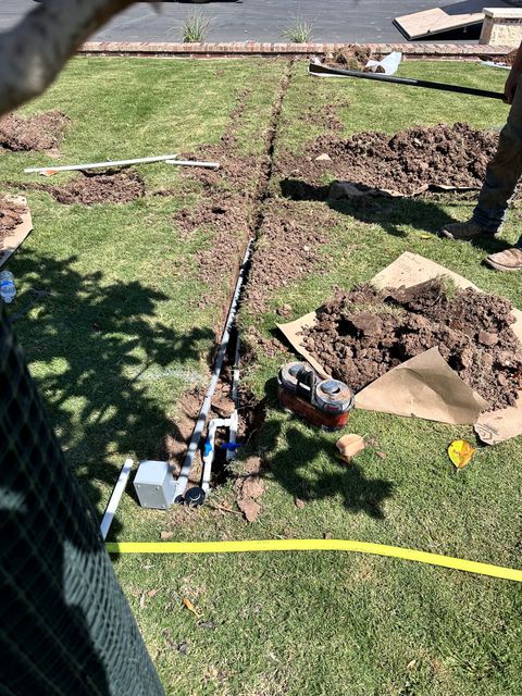Trench dug in grass with irrigation system parts. Soil piles, yellow hose, blue valves, and a person’s legs are visible.