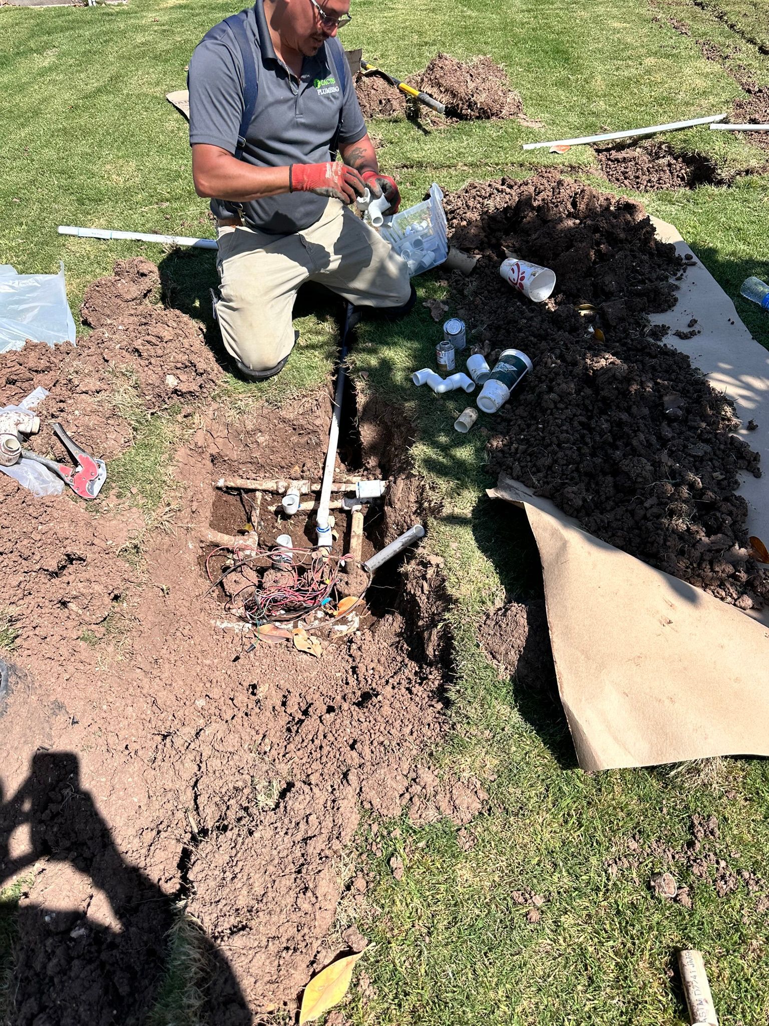 Man kneeling, repairing irrigation system in a yard; pipes and tools visible, sunny outdoors.