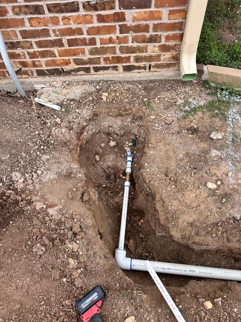 PVC pipe and water valve in a dirt trench near a brick wall.
