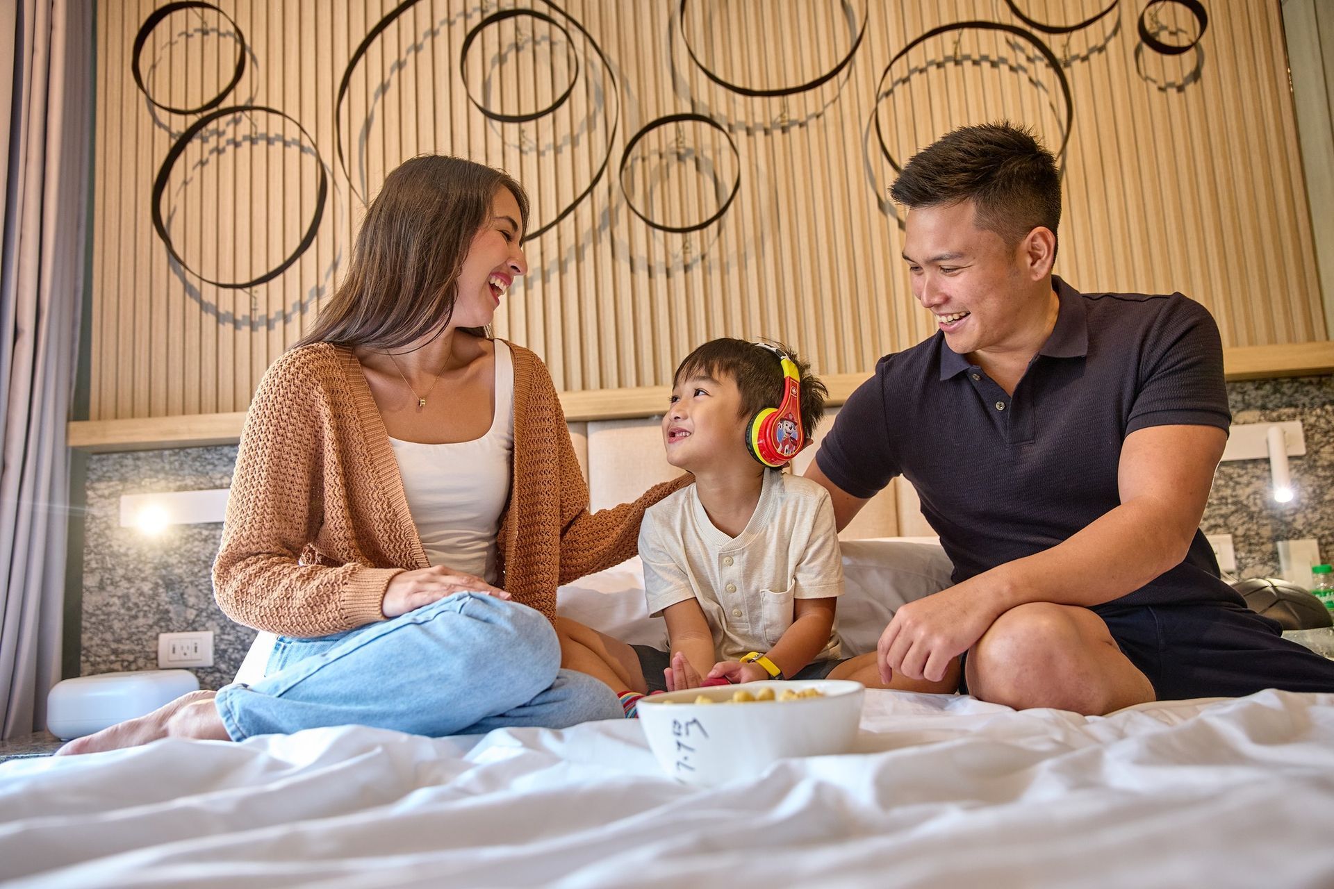 Three people sitting on a bed, smiling and talking in a cozy bedroom with circular wall decor.
