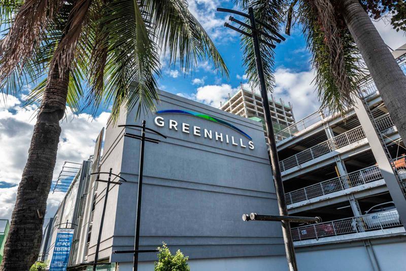 Greenhills mall entrance with palm trees and blue sky beside a multi-level parking structure