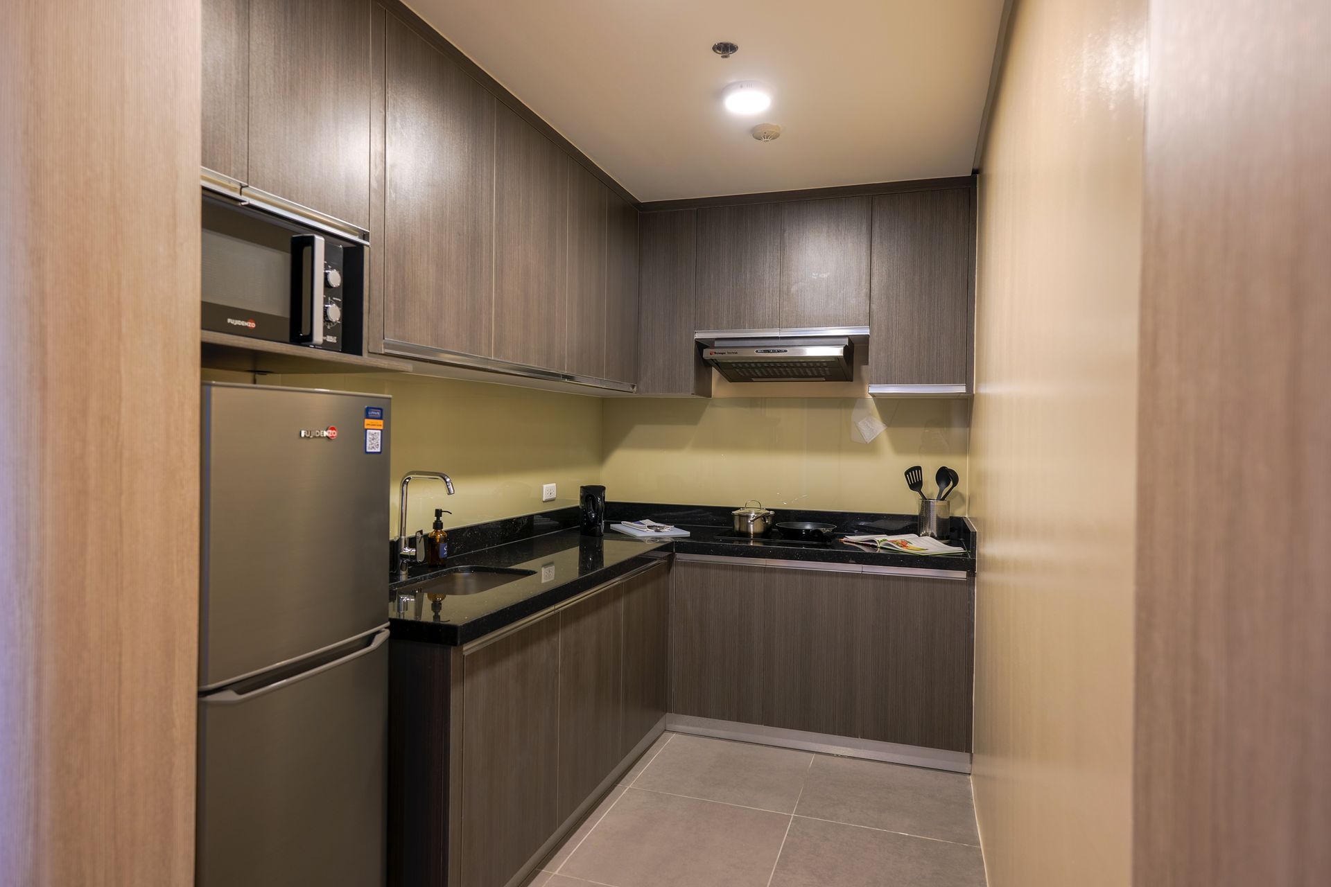 Compact kitchen with wood cabinets, stainless fridge, black countertops, and tiled floor.