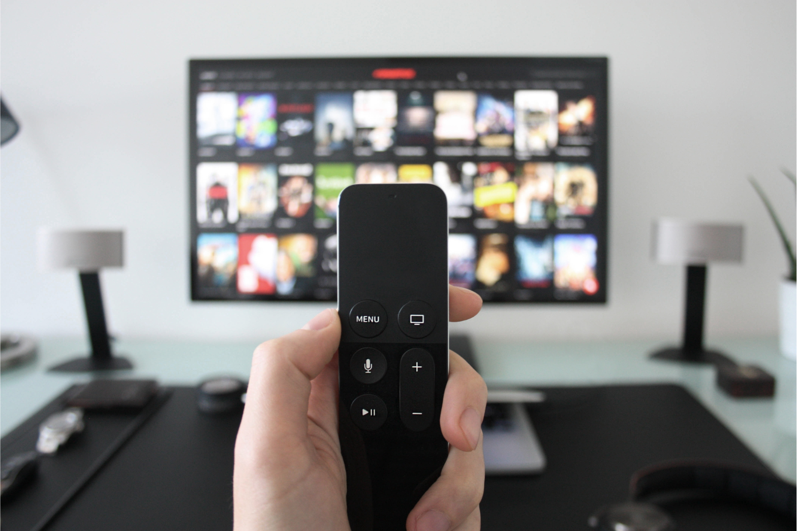 Hand holding a black remote control in front of a TV screen with a streaming service menu.
