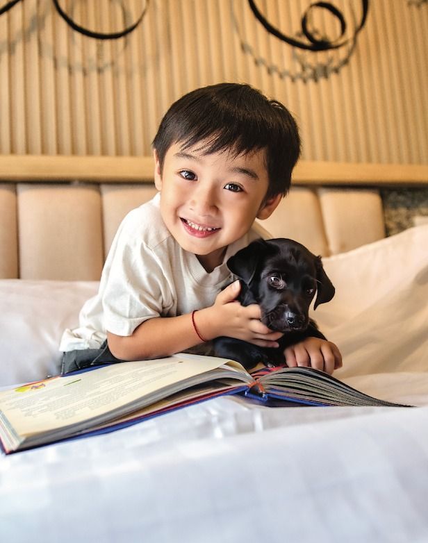 Boy cuddling a black puppy on a bed, smiling at the camera, book open in front of them.