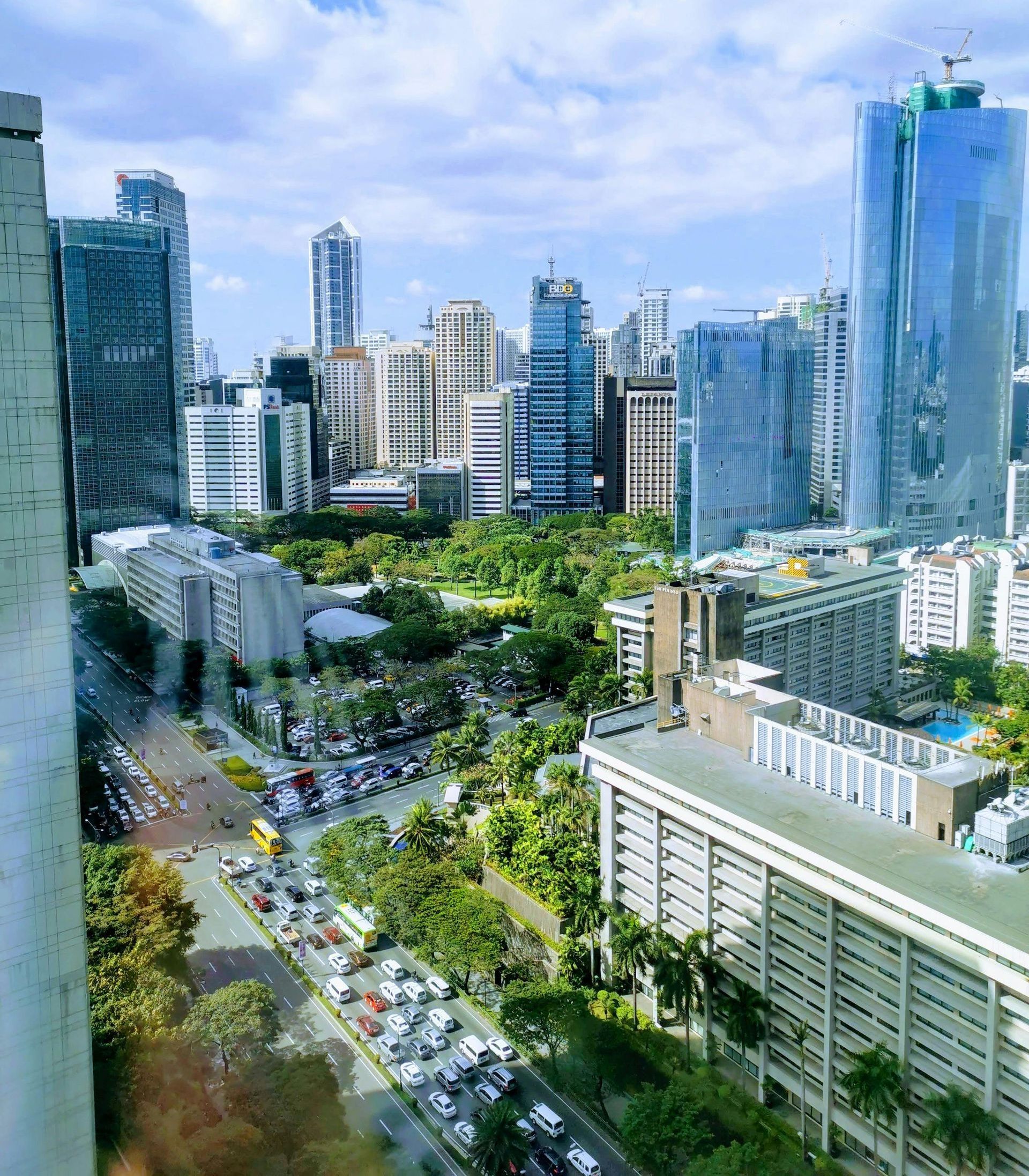 Cityscape view with skyscrapers, green trees, a park, and cars on the road under a blue sky.