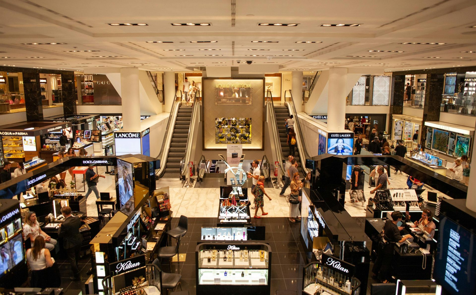 Crowded multi-level department store interior with escalators, bright lights, and cosmetics counters