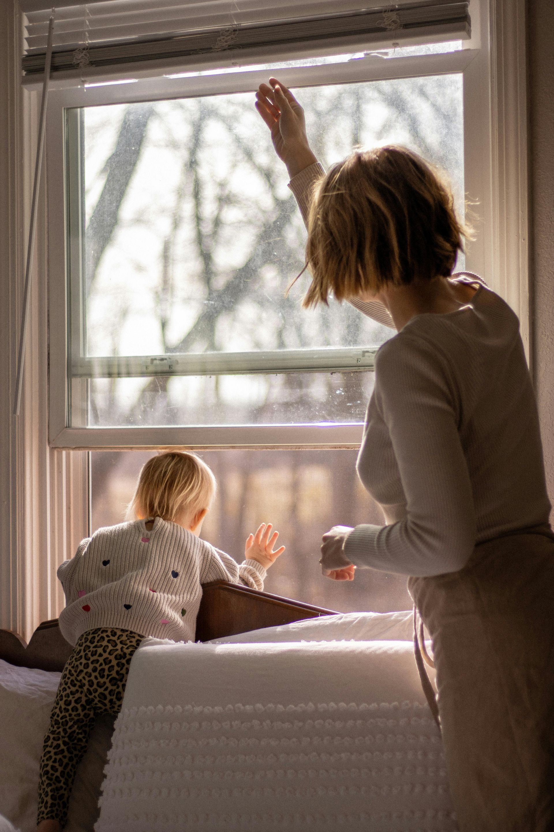 Mother and toddler looking out a sunlit bedroom window together indoors