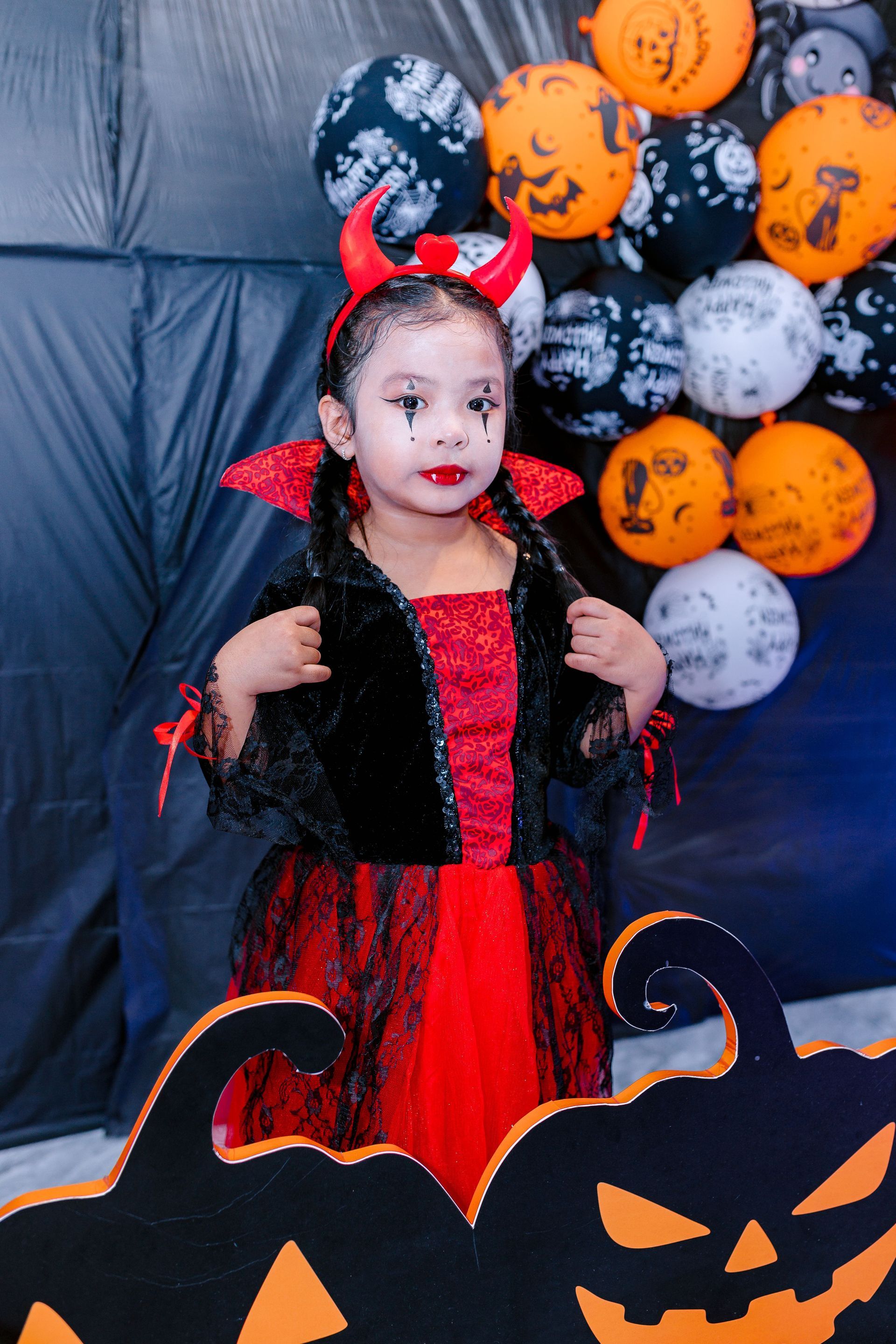 Child in a red devil costume with horns and wings, standing before Halloween balloons and a jack-o'-lantern display