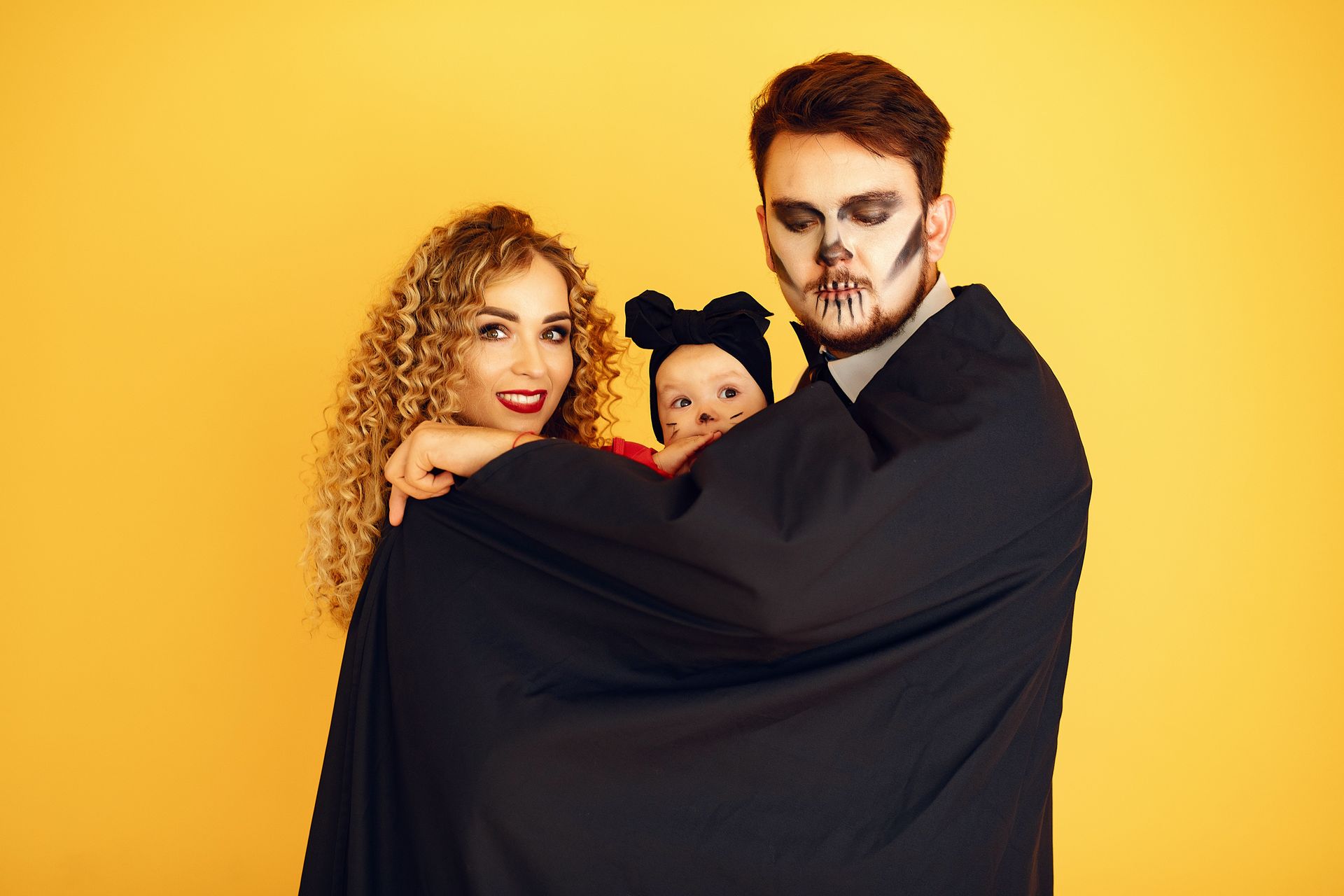 Three people wrapped in a black cloak, posing against a bright yellow background.
