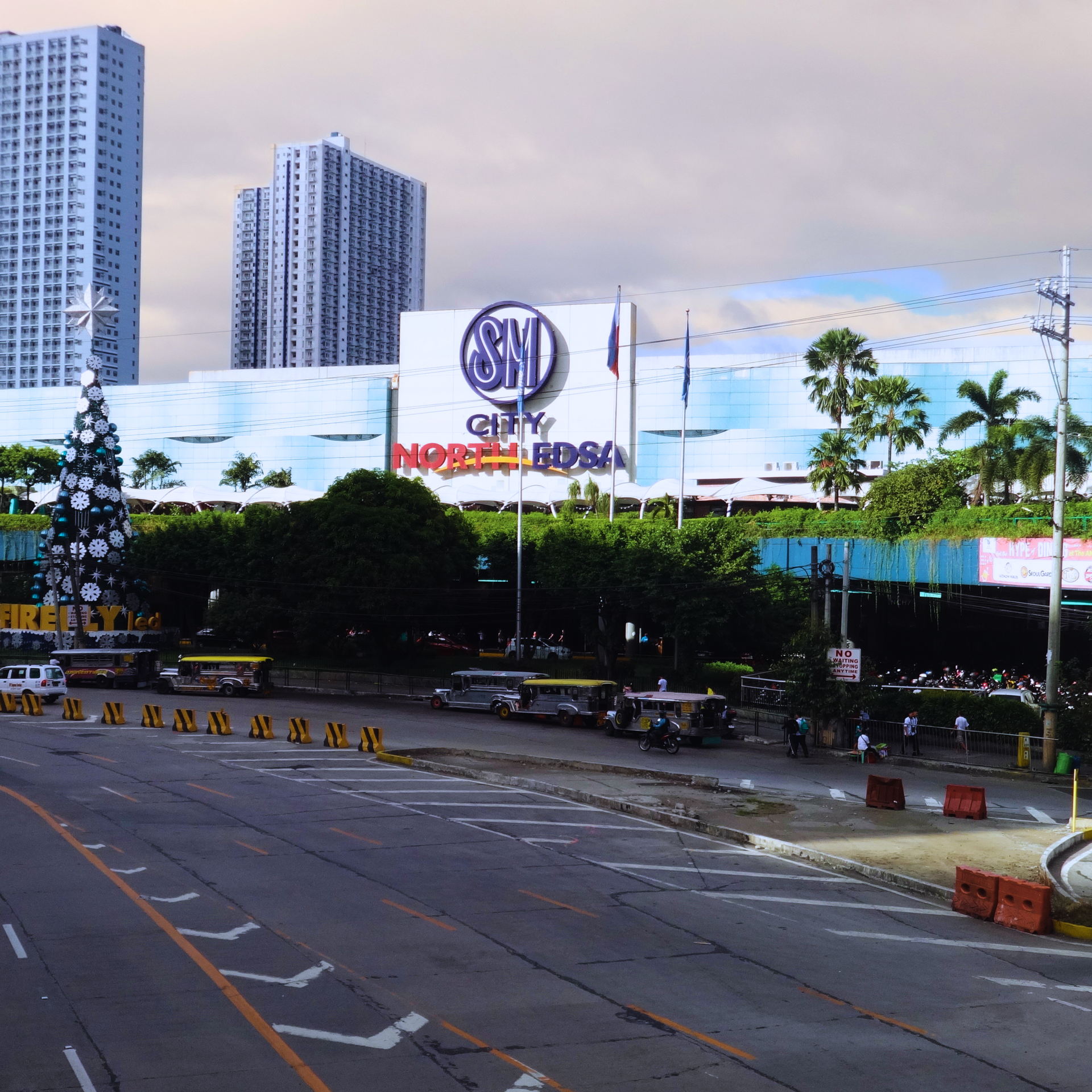 SM City North EDSA with tall buildings in the background, a street with vehicles, and a Christmas tree.