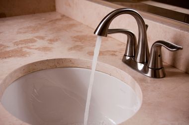 A stainless steel faucet running water into a white bathroom sink set in a beige stone countertop.