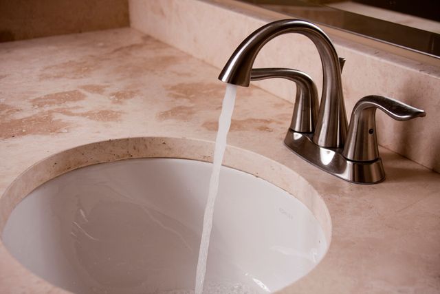 A chrome kitchen faucet sits on a dark stone countertop next to a circular stainless steel sink with marble-pattern walls.