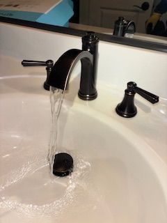 A dark metal bathroom faucet pours water directly into the center of a white ceramic sink basin.